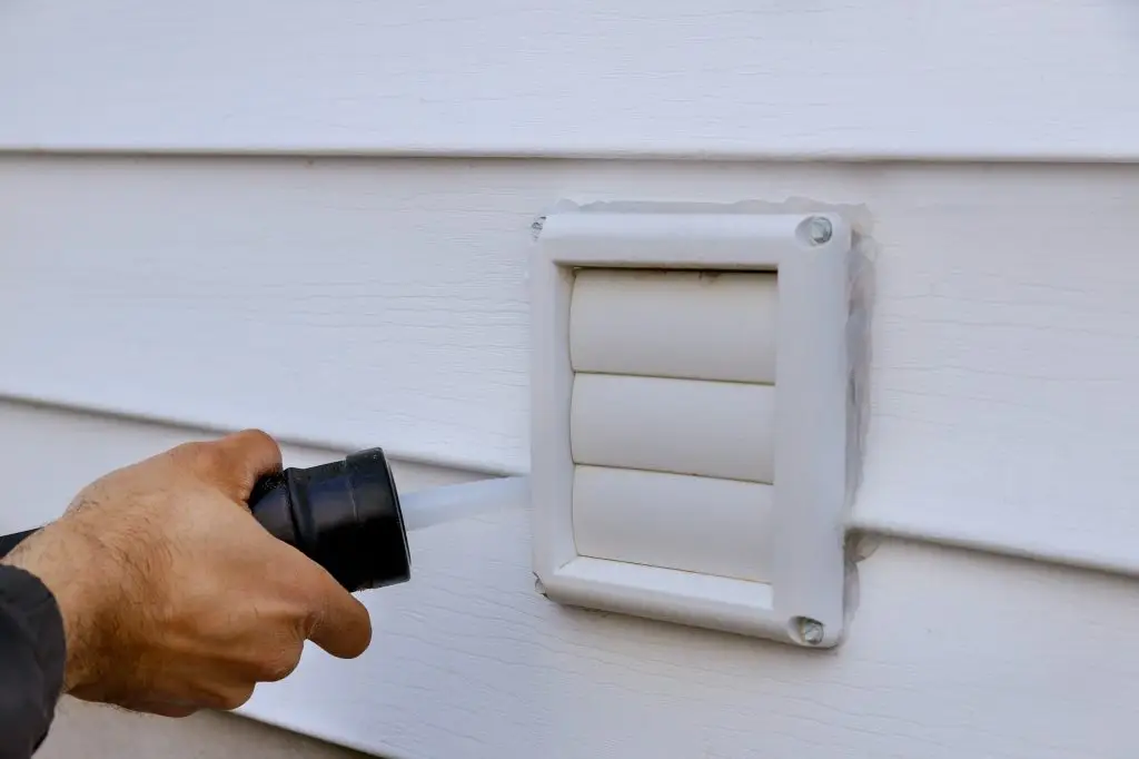 Applying silicone on plastic exhaust fan on the wall outside siding trim