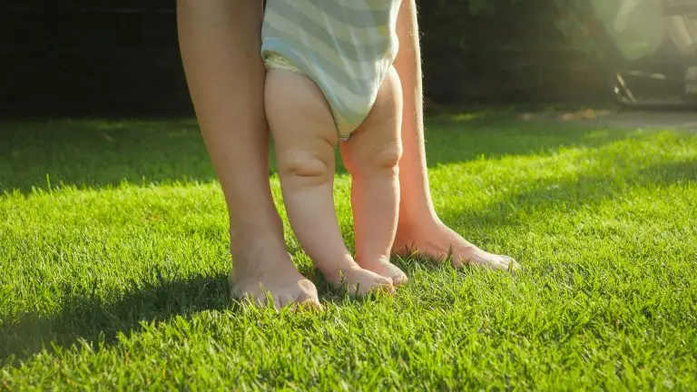 Closeup of barefoot baby with mother standing on fresh green grass lawn at house backyard garden