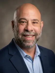 A smiling bald man, Franklin Hernandez, with a salt-and-pepper beard wearing a blue suit jacket and light blue shirt, poses in front of a neutral, blurred background.